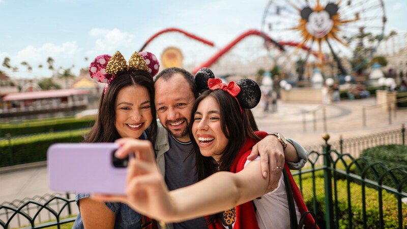 A young woman taking a selfie with her mom and dad in front of Pixar Pier at Disney California Adventure Park