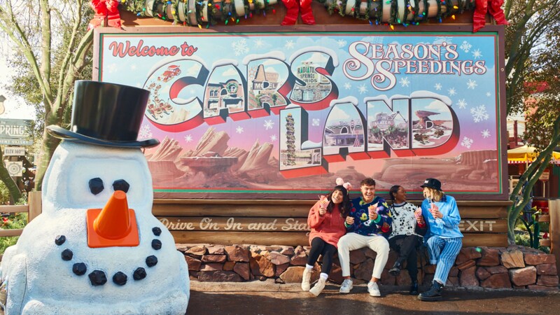 Mater made up as a snowman and young people enjoying snacks in front of a billboard reading ‘Welcome to Cars Land, Season’s Speedings’