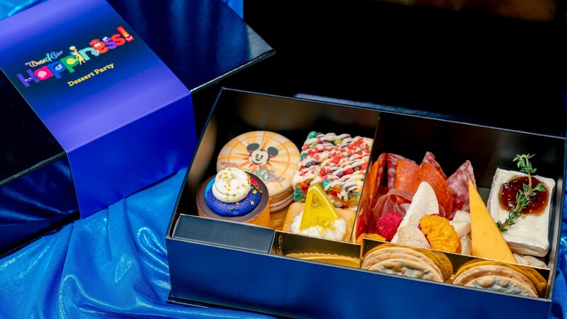 An open box of assorted sweets and charcuterie items with a message on the box lid reading World of Color Happiness Dessert Party