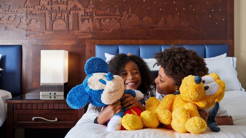 Two children smile and cuddle Mickey Mouse and Pluto plush toys while relaxing on a hotel bed at the Disneyland Hotel