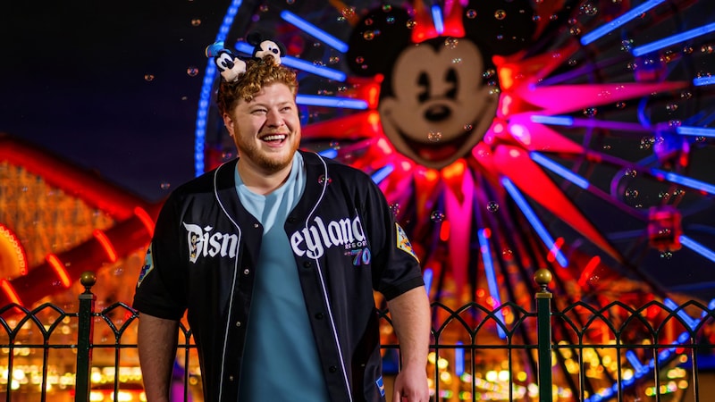 Guest wearing a Disneyland 70th anniversary baseball jersey and Mickey Mouse ear headband stands near a railing with a brightly lit Ferris wheel featuring a Mickey face behind them at night