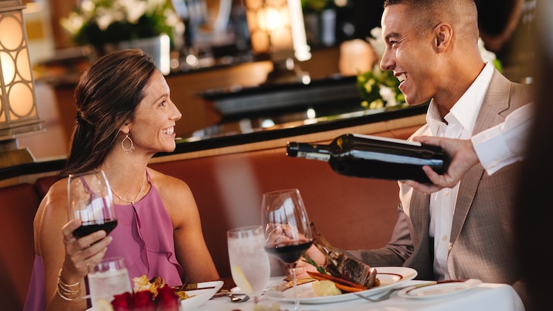 A man and a woman smiling at each other over dinner while a server pours wine into a stemmed glass
