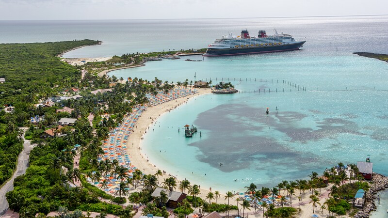 A Disney ship docked next to a tropical beach resort with palm trees, umbrellas and lounge chairs