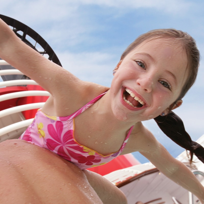 A small girl smiling with joy as her father carries her through the pool area of a Disney cruise ship