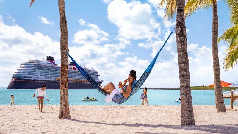 A woman in a hammock between palm trees and other Guests relaxing on a beach with a Disney Cruise Line ship off the coast