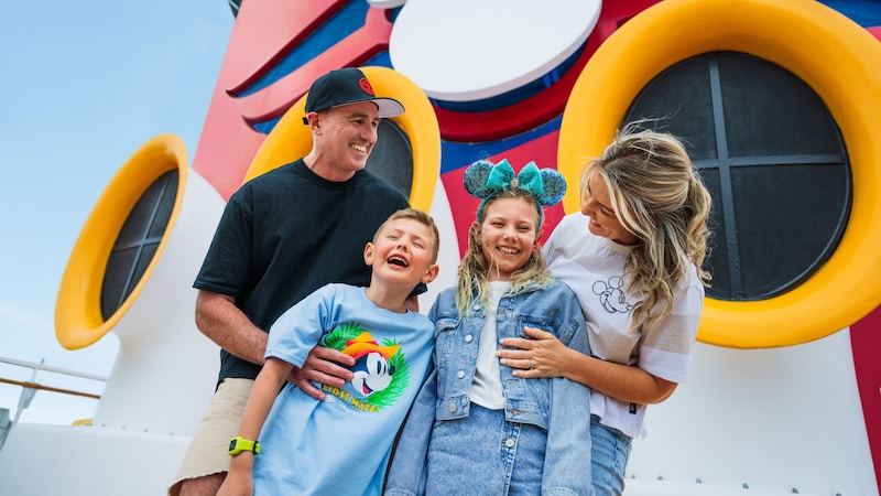 Family of 4 smiling in front of a Disney Cruise Line ship funnel Family of 4 smiling in front of a Disney Cruise Line ship funnel