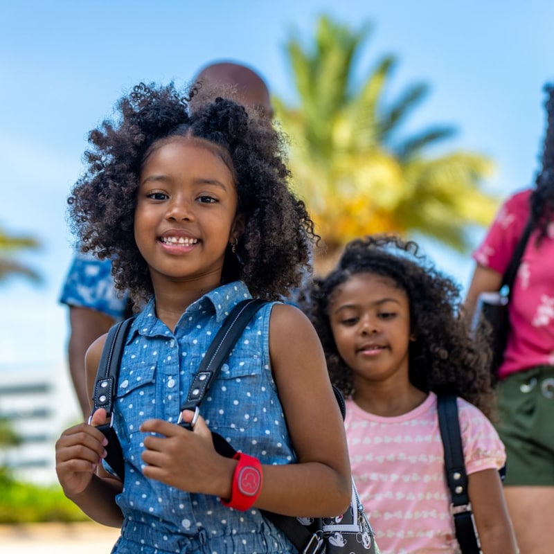 A young girl wearing a Disney Band Plus smiles while walking with her younger sister and parents in a tropical setting