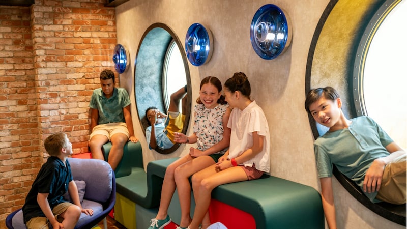 Edge A group of 6 tweens sitting and interacting in a play area with large portholes, modern seating and brick walls