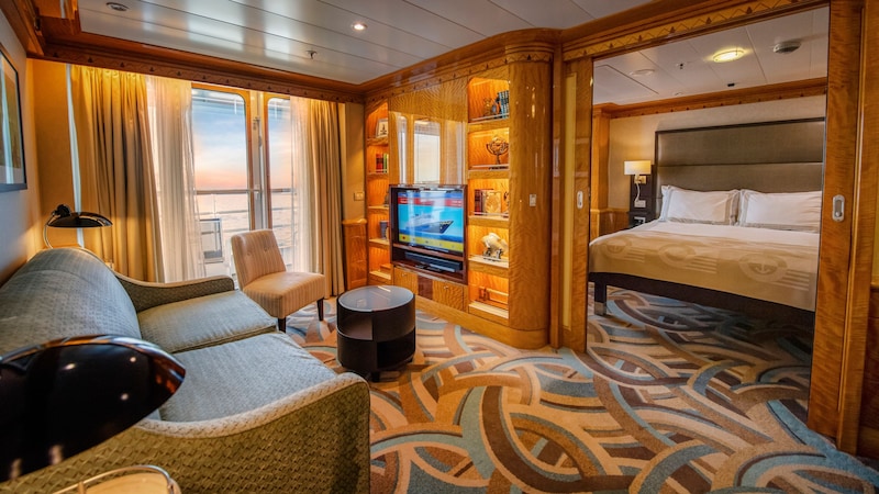 Concierge 1-Bedroom Suite with Verandah The interior of a stateroom suite reveals a living area with a private balcony, sofa, chair, coffee table, built in bookcase, TV and a sliding door that leads to a bedroom with a large bed