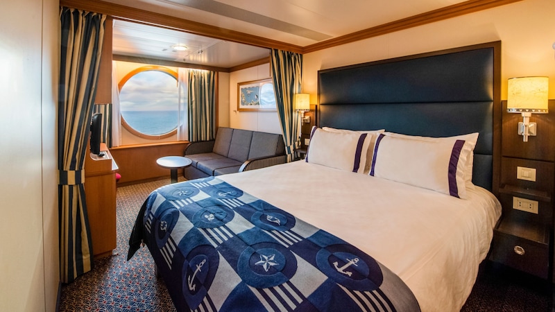 Deluxe Oceanview Stateroom A stateroom with a queen bed, privacy curtain, sofa, coffee table, shelves and a porthole window