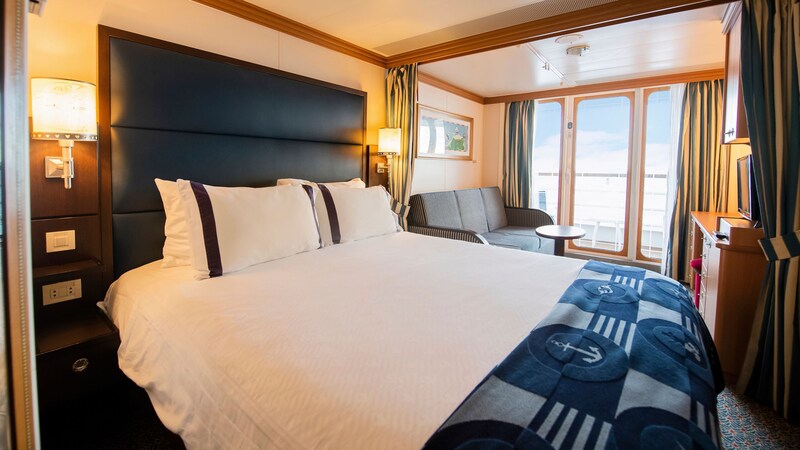 A stateroom with a queen bed, privacy curtain, sofa, coffee table, shelves and a private balcony
