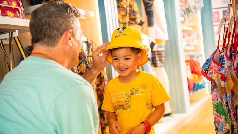 A smiling boy trying on a Disney Cruise Line bucket hat in a gift shop aboard a Disney cruise ship
