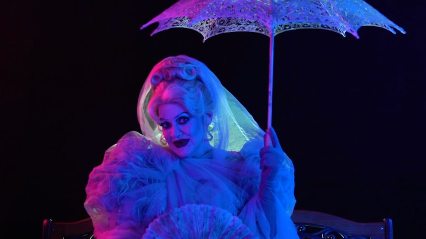 Miss Carlotta, decked out in her last earthly garments and holding a hand fan and an umbrella, sits on a bench as she smiles an other worldly smile