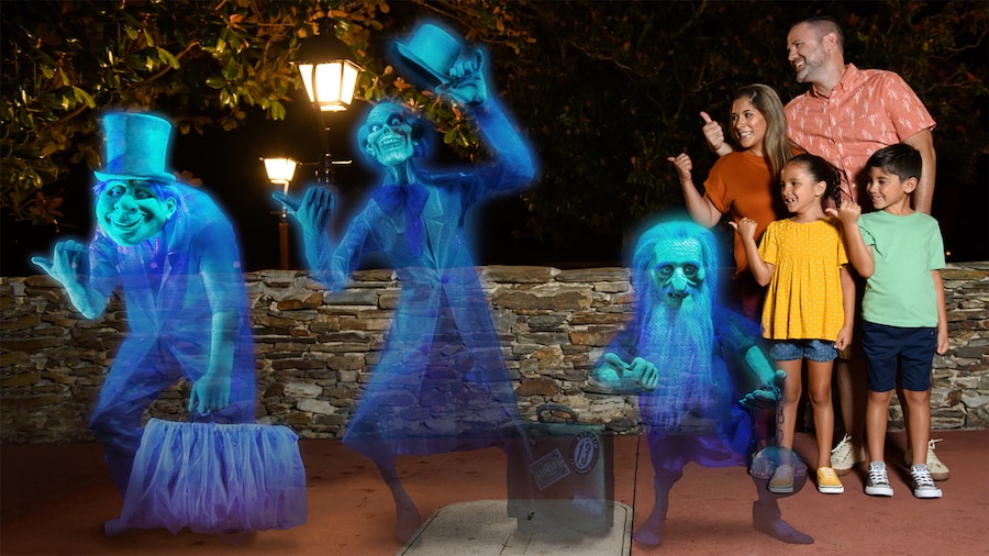 A mom, dad and their two kids give a hitchhiking thumbs up while standing near the three grim grinning ghosts from the Haunted Mansion at night