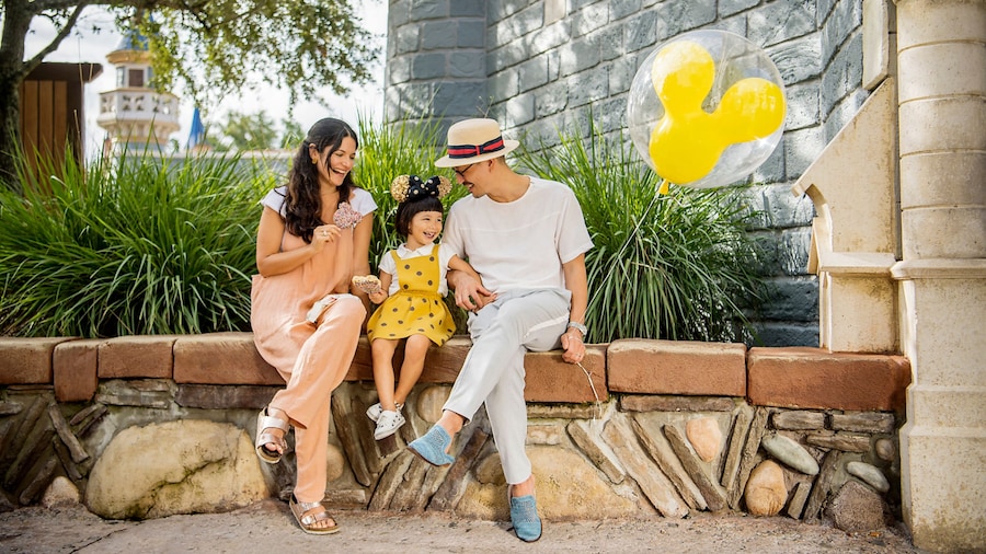 A man, woman and their daughter sitting on a bench with a Mickey Mouse shaped balloon in Magic Kingdom park