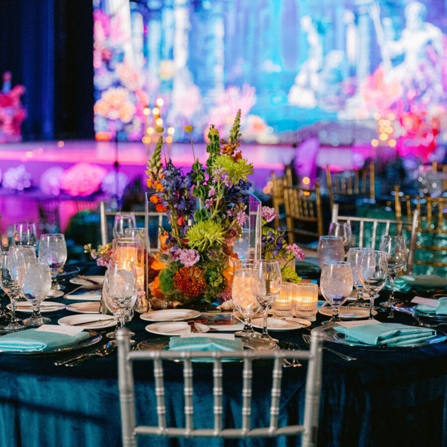 Un lugar para eventos con salón de baile, escenario, mesas de comedor, sillas, arreglos florales y cubiertos