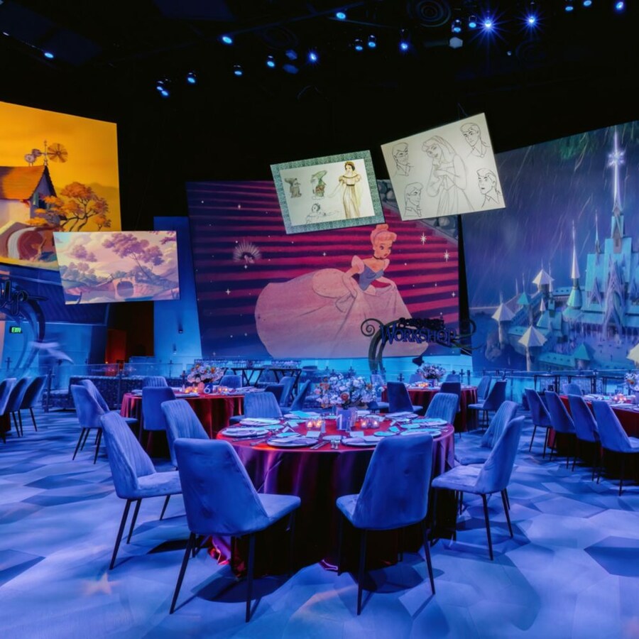 Un espacio para eventos con salón de baile con mesas de comedor, sillas, arreglos florales, cubiertos y pantallas que muestran obras de arte de películas de Disney.