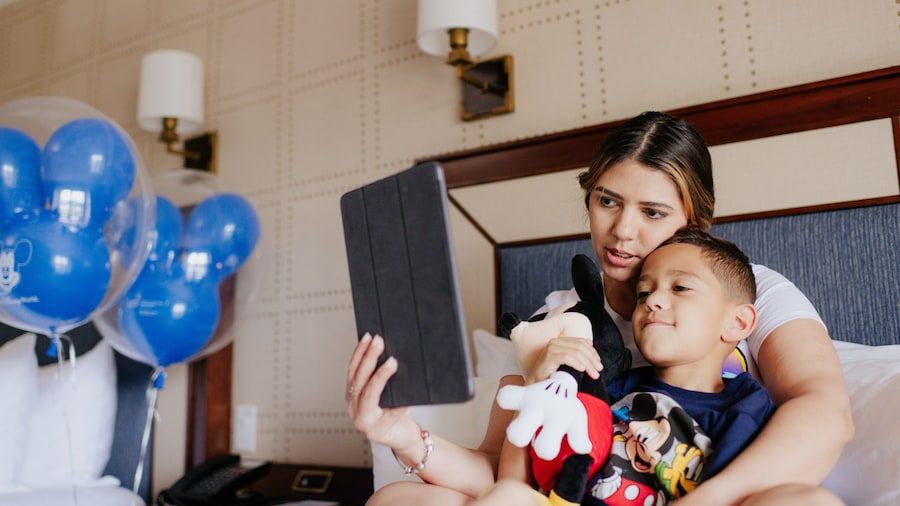 A woman sits on a bed in a hotel room and shows an I Pad screen to her son, who sits with her