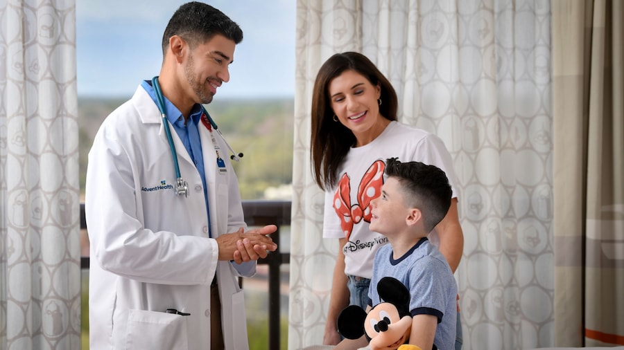 A doctor talking to a boy sitting on a bed with a Mickey Mouse plush while his mother stands next to him