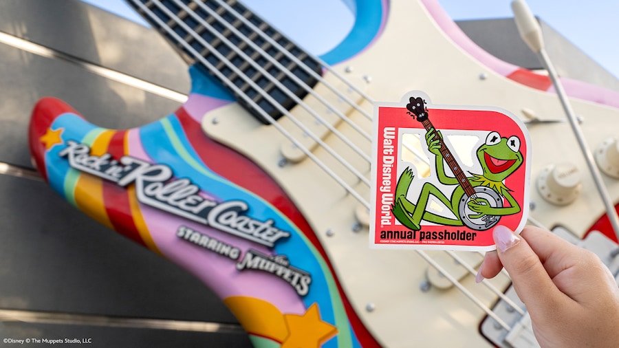 A hand holding a Walt Disney World Annual Passholder magnet featuring Kermit the Frog playing a banjo in front of the entrance to Rock n Roller Coaster at Disney's Hollywood Studios