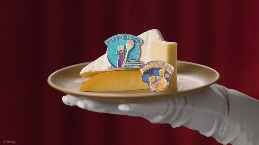 A gloved hand holding a plate of cheese and Epcot International Food & Wine Festival 2025 Passholder trading pins featuring Remy and a plate of food