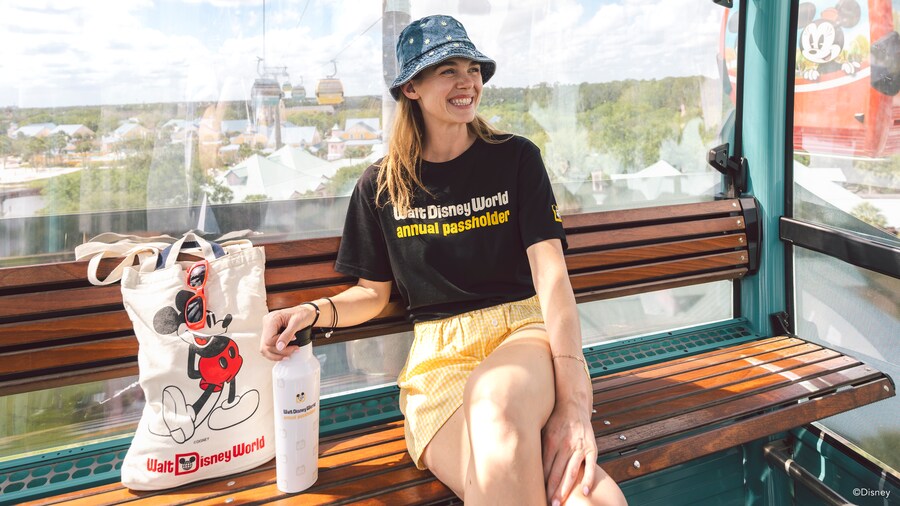 A seated woman wearing a Walt Disney World Annual Passholder T-shirt with an Annual Passholder water bottle and a tote bag featuring Mickey Mouse