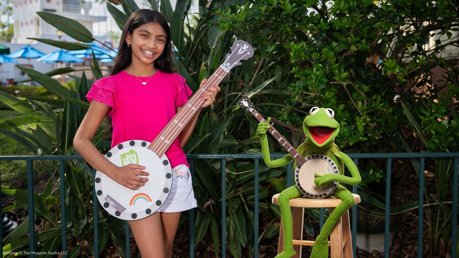 A young girl smiling while holding a painted banjo next to Kermit the Frog playing a banjo