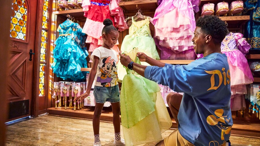 A man holding up a Princess Tiana dress in front of a girl at Bibbidi Bobbidi Boutique