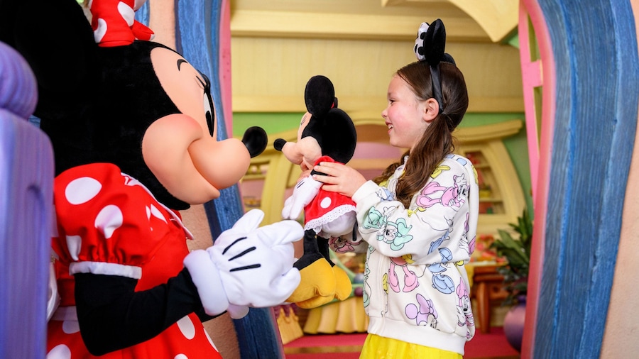 A girl smiling and holding up a plush Minnie Mouse in front of Minnie Mouse