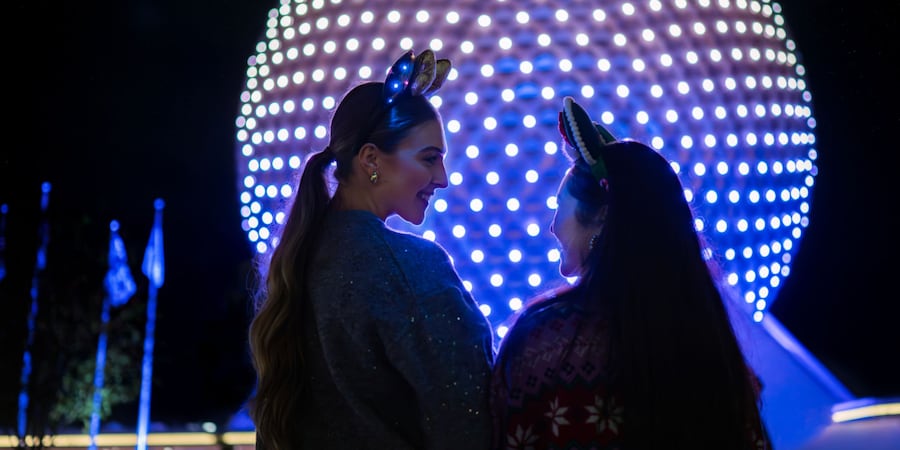 Dos mujeres jóvenes con diademas de orejas de Minnie Mouse se sonríen entre sí frente a Spaceship Earth en Epcot