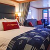 Stateroom with bed, dividing curtain, sofa, glass door that opens to verandah, built in wooden desk and TV