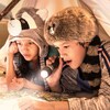 Two kids wearing Frontierland inspired novelty hats read a map with a flashlight and a small lantern underneath a makeshift teepee on their Resort bed