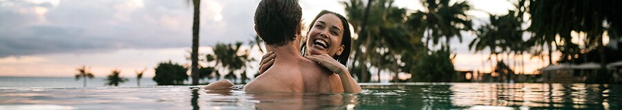 Two smiling adults in an infinity pool, with palms silhouetted along the shoreline