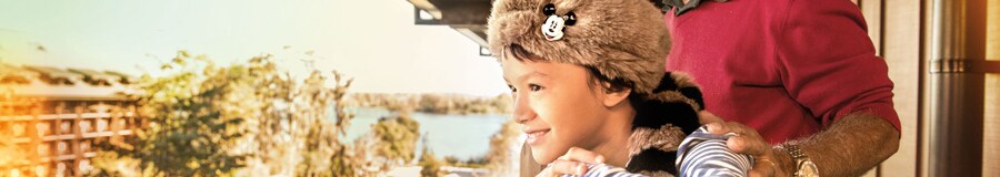 A young boy wearing a novelty coonskin hat takes in views from their room’s balcony with his father
