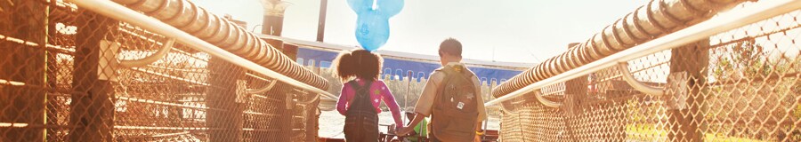 A young sister with a Mickey ears balloon holds her brother’s hand while crossing a bridge