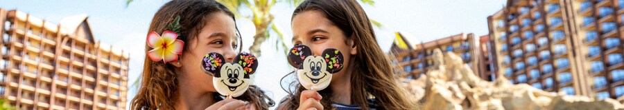Two little girls look at each other as they eat their Mickey Mouse lollipops