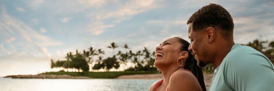 Two Guests sitting together on the beach while the sun sets over Aulani, A Disney Resort & Spa