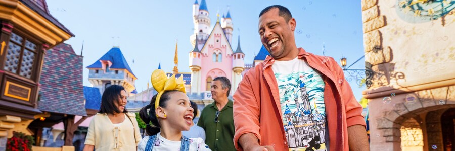 A young girl wearing a Minnie Mouse headband walking with her dad through Disneyland Park with Sleeping Beauty Castle in the background