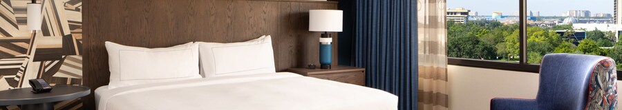 A room with a small table, a king bed, a nightstand, a cushioned chair, a cabinet and a window with exterior views at the DoubleTree Suites by Hilton Orlando