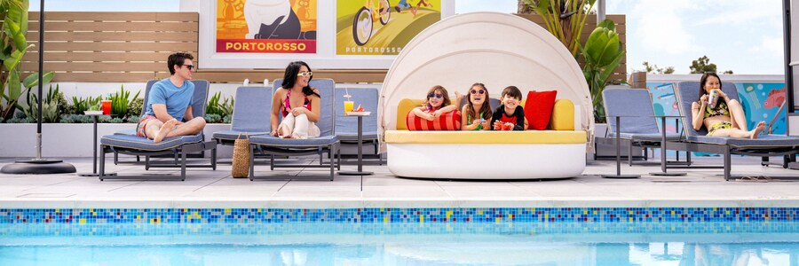 At the rooftop Pixel Pool area at Pixar Place Hotel, adults sitting in lounge chairs and 3 kids relaxing on a day bed, surrounded by vibrant artwork depicting Pixar stories