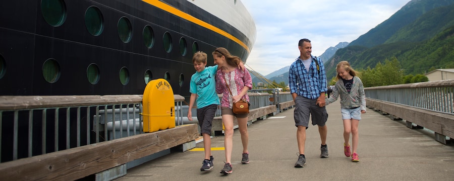 A young family of 4 walking alongside a docked Disney cruise ship in a mountainous setting A young family of 4 walking alongside a docked Disney cruise ship in a mountainous setting