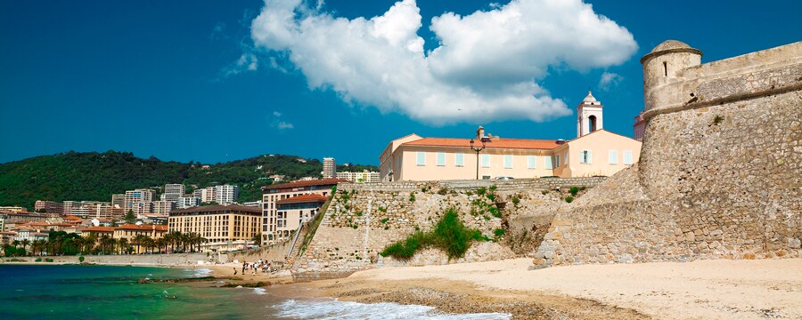 People on a beach near a sea wall, buildings, trees and mountains