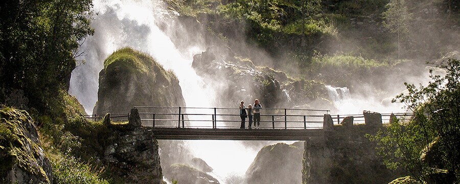 Three people stand on a bridge looking out with a waterfall cascades down rocks behind them