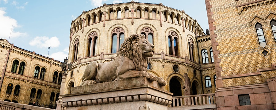 Statue of a lion sits infront of a circular building with the Norwiegan flag on its roof Statue of a lion sits infront of a circular building with the Norwiegan flag on its roof