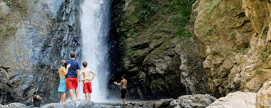 People stand in front of a tall waterfall pouring down a rock face People stand in front of a tall waterfall pouring down a rock face
