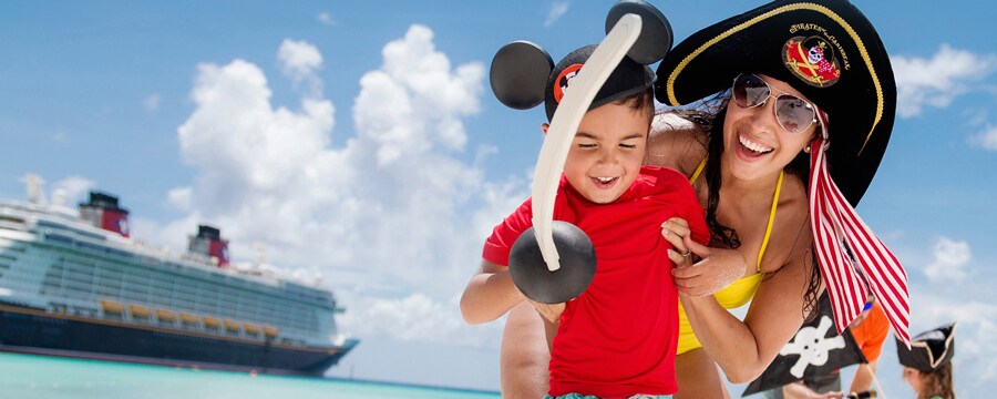 With a Disney cruise ship in the background, a woman in a swimsuit and pirate’s hat smiles while playing with her small son, who is wearing Mickey ears and holding a plastic pirate’s sword