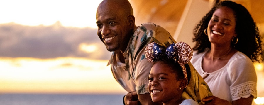 A smiling girl, wearing Mickey ears, stands on the deck of a ship with her parents as they all look out to sea A smiling girl, wearing Mickey ears, stands on the deck of a ship with her parents as they all look out to sea