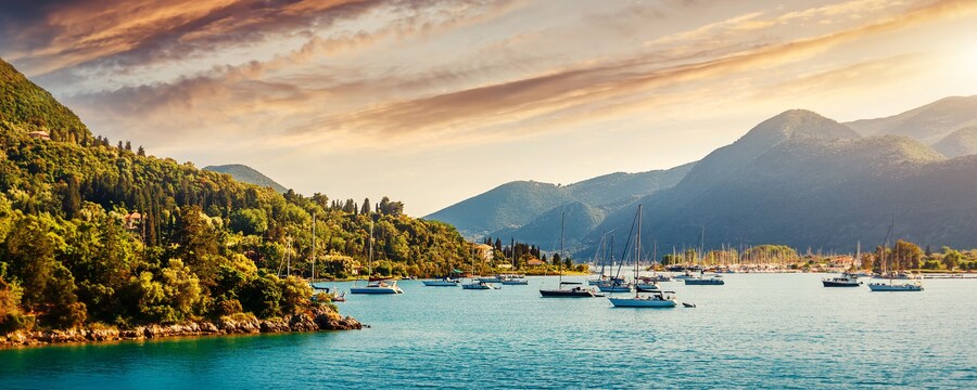 A bay in Corfu with sailboats anchored near a lush shoreline and mountains in the distance