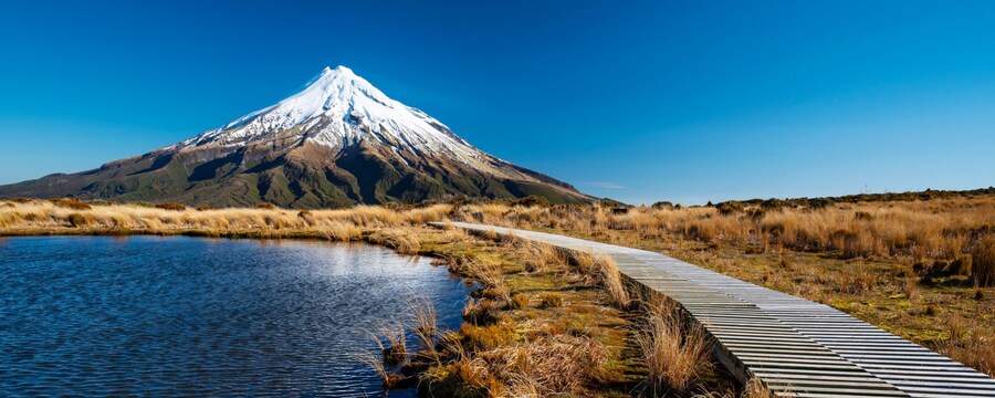 A boardwalk next to a lake with Mount Taranaki in the distance