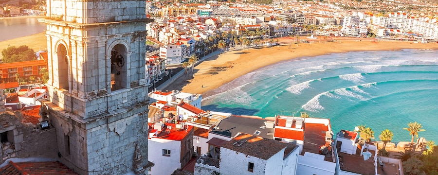 A coastal town featuring a stone belltower on a hill overlooking a beach A coastal town featuring a stone belltower on a hill overlooking a beach